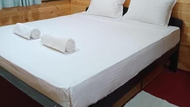 A single bed with white bedding and two rolled towels, a wooden wall, and a red carpet. - Hiriketiya Budget Stay (Dikwella)