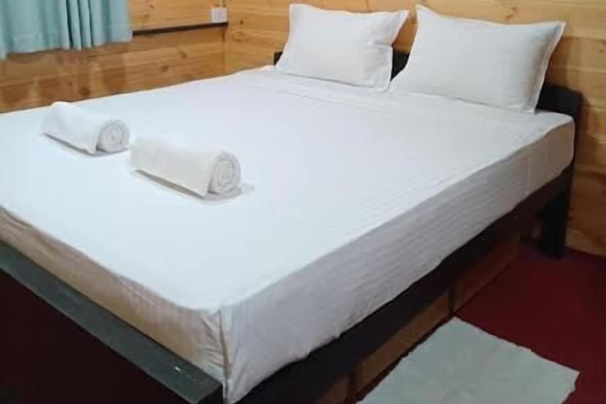 Un lit simple avec une literie blanche et deux serviettes enroulées, un mur en bois et une moquette rouge.