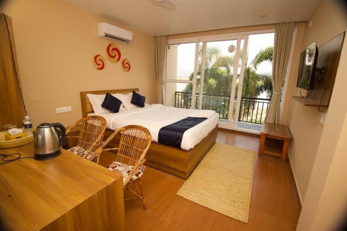 A hotel room with a bed, a desk, a chair, a TV, and a balcony with a view of palm trees.