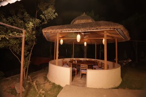 A circular outdoor dining area with a thatched roof, wooden tables and chairs, and hanging light fixtures.