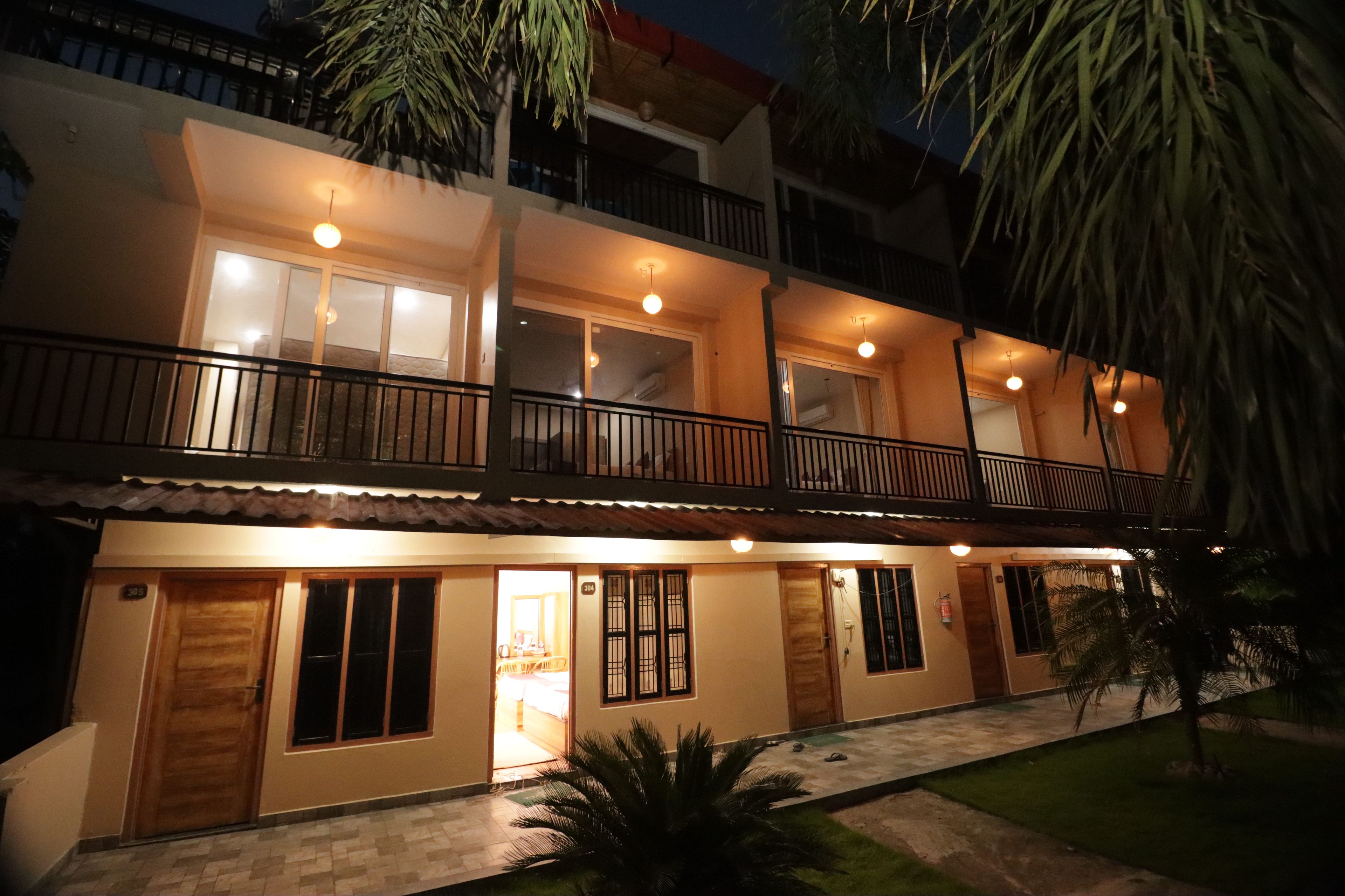 A two-story building with a balcony on the upper floor, a door with a window on the ground floor, and a small garden area with a palm tree.