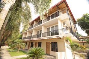 A multi-story building with balconies, a flat roof, and a surrounding garden with palm trees.