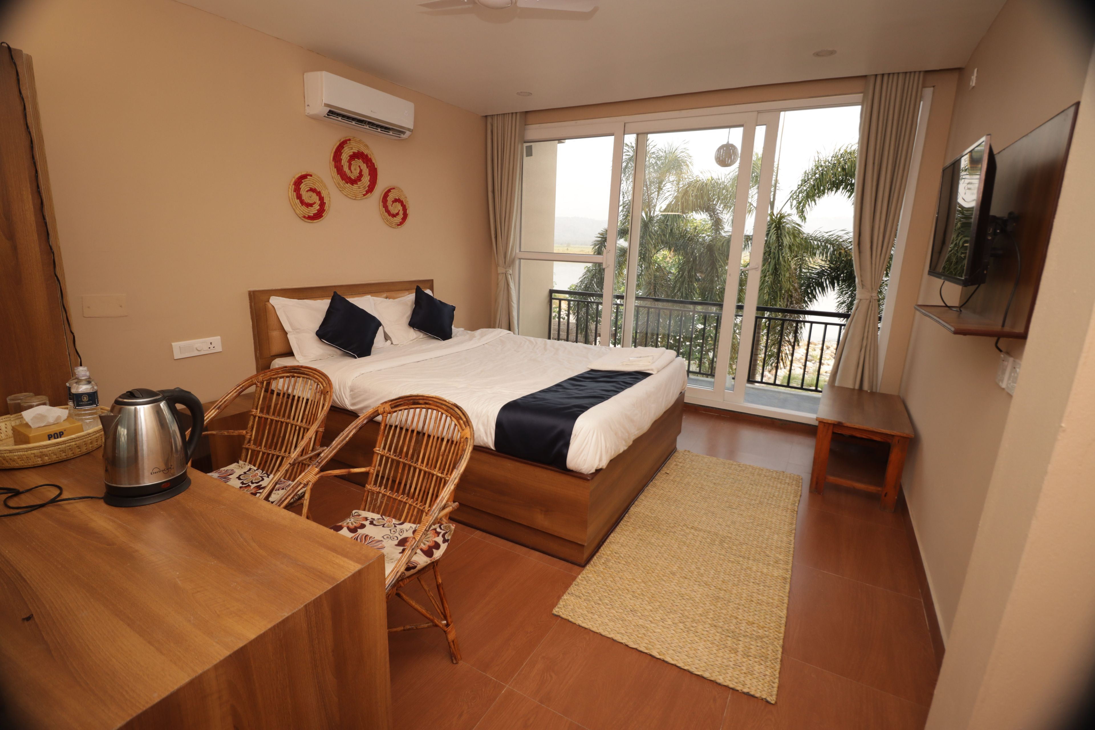 A hotel room with a bed, two chairs, a table with a kettle, a TV, and a balcony with a view.