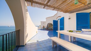 Una terraza con vista al mar, que incluye una mesa con un cuenco de fruta, contraventanas azules y techo de paja.