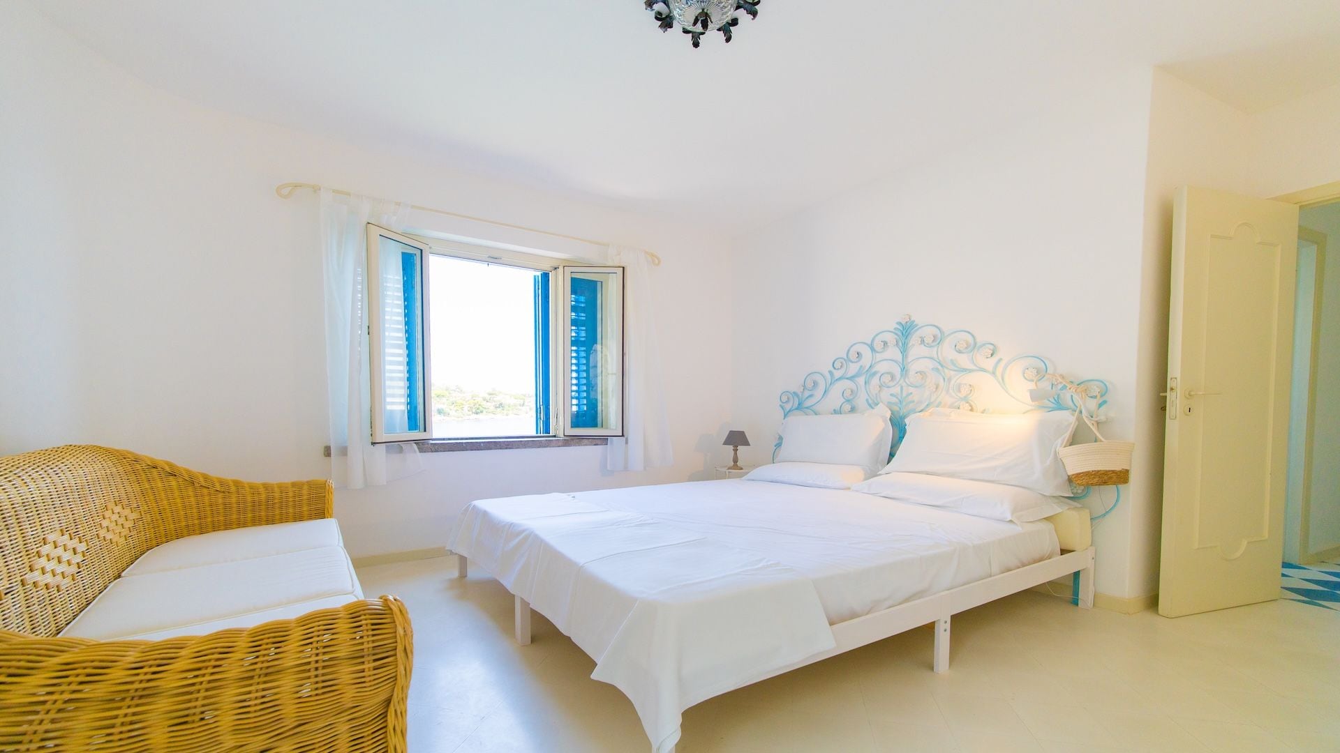 Quarto com cama, mesa de cabeceira, abajur, cadeira de vime e janela com cortinas.
