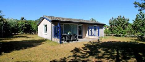 A small house with a single door and window, a patio with a table and chairs, and a fenced yard.
