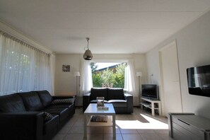 A living room with a sofa, a coffee table, a television, and a window with curtains. - Bungalow in Ballum with swimming pool (Ballum)