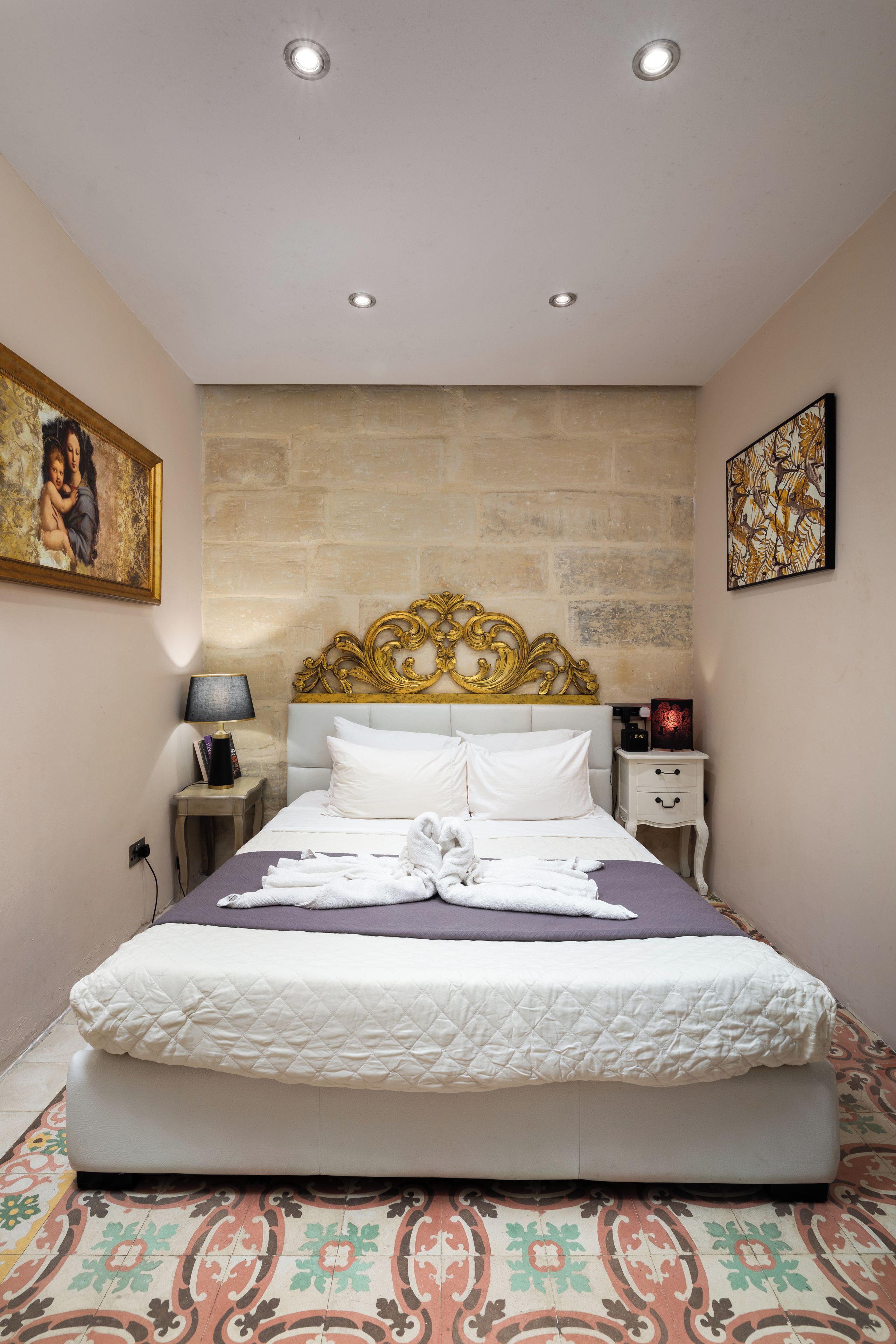 A hotel room with a bed, bedside tables, a nightstand, a lamp, a painting, and a decorative headboard.