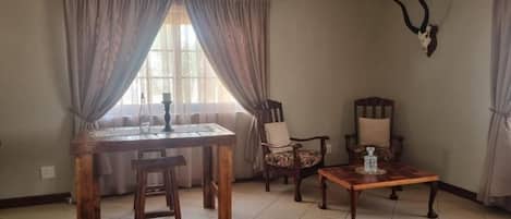 A dining area with a wooden table, two chairs, a bench, and a coffee table. There is a window with curtains, a wall-mounted antler decoration, and a tiled floor.