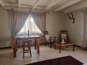 A dining area with a wooden table, two chairs, a bench, and a coffee table. There is a window with curtains, a wall-mounted antler decoration, and a tiled floor. - Skilpadvlei Accommodation (Velddrif)