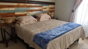 Une chambre à coucher avec une tête de lit en bois, un lit avec une literie beige, un plaid bleu et un petit panier en osier posé sur une table de chevet.