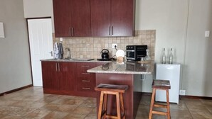 Una cocina con armarios de madera oscura, salpicadero de azulejos y una encimera con hervidor, cafetera y dos taburetes.