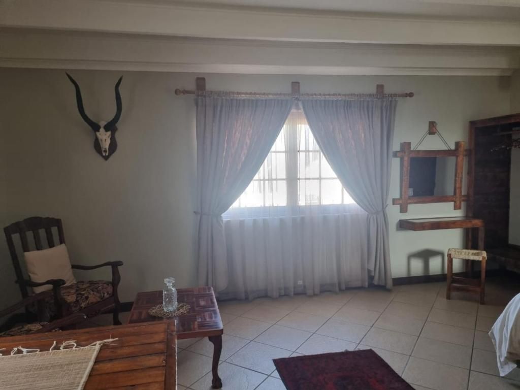 A room with a large window, curtains, a wooden chair, a small table with a bottle, and a wall-mounted deer skull.