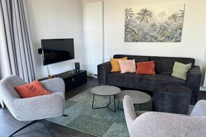 Ein modernes Wohnzimmer mit einem grauen Sofa, zwei Sesseln, einem Couchtisch und einem an der Wand befestigten Flachbildfernseher.