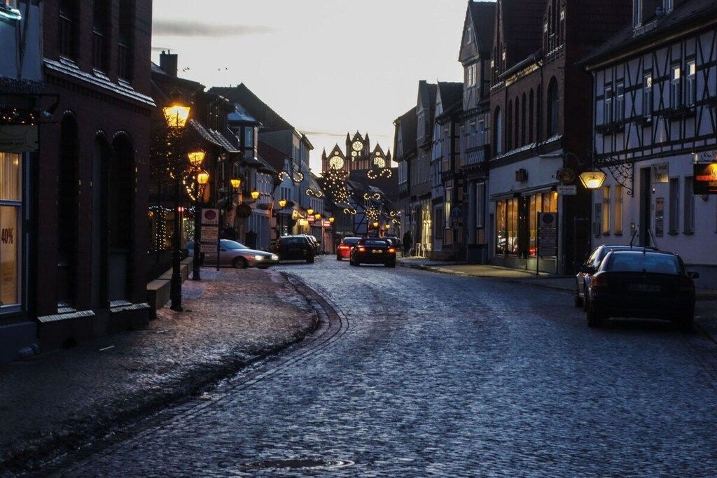 A cobblestone street lined with traditional European buildings, some with illuminated signs and decorations.