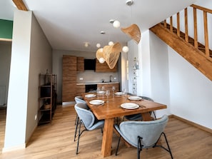 A modern dining area with a wooden table set for four, surrounded by chairs, and a kitchen in the background. - Vrbo Property (Chartres)