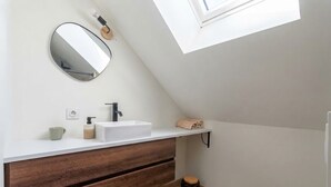 Moderni kylpyhuone, jossa on kattovalokuvio, valkoinen allaskaappi, puiset laatikot, pyöreä peili ja wc-istuin.