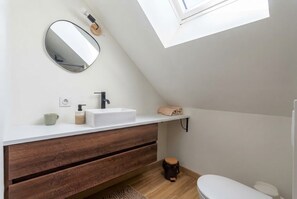A modern bathroom with a skylight, white vanity, wooden drawers, a round mirror, and a toilet. - Vrbo Property (Chartres)