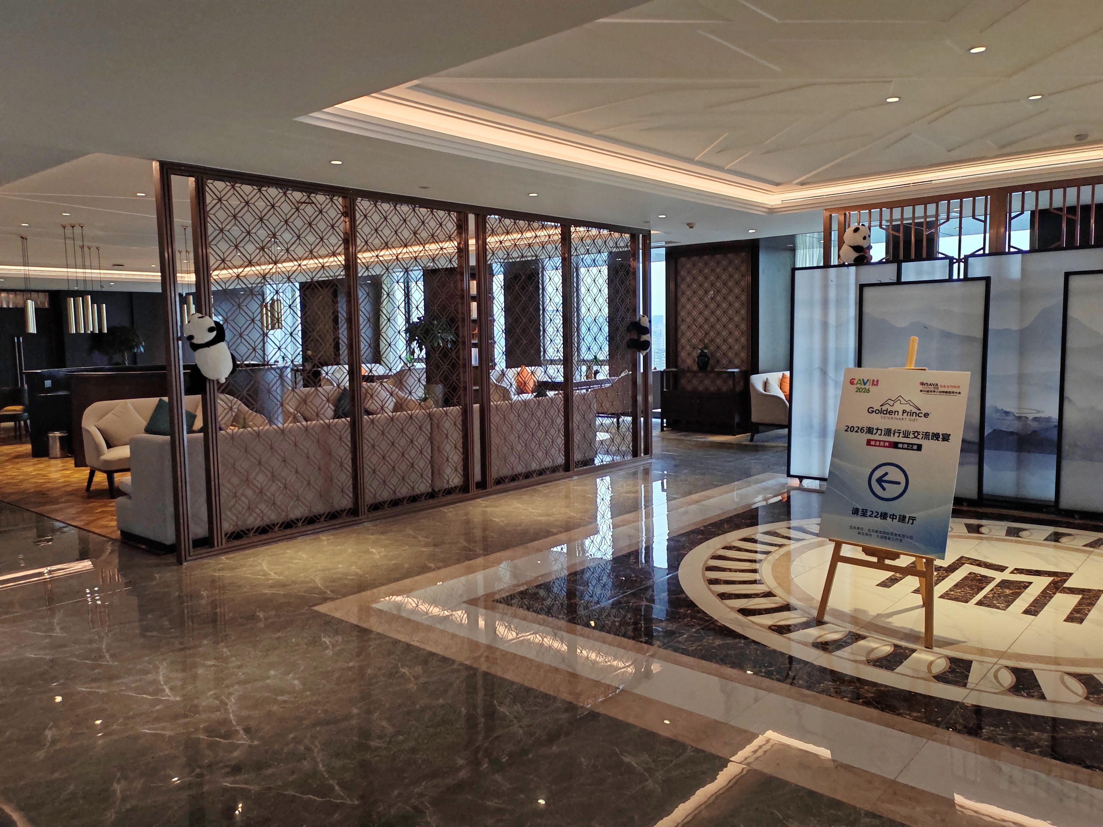 A modern hotel lobby with a seating area, a reception desk, and a sign indicating a special event.