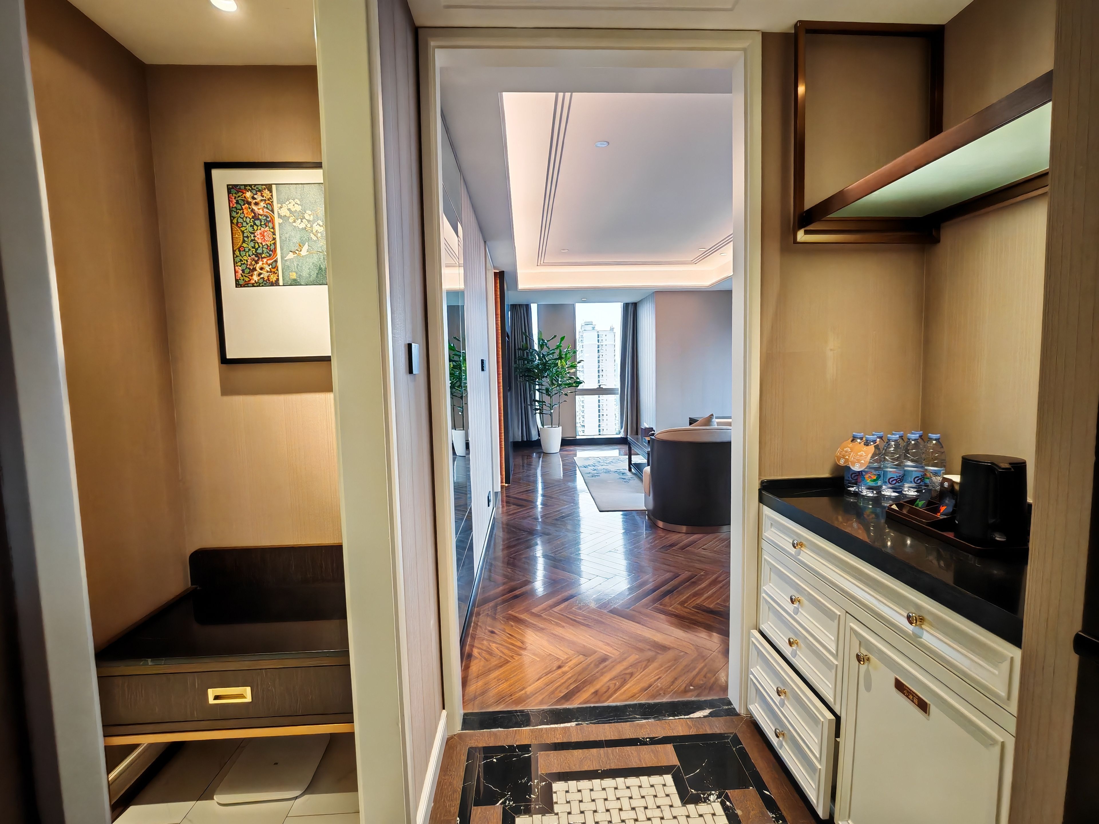 A hotel corridor with a wooden floor, a seating area, a kitchen with a coffee machine, and a view of the cityscape through the windows.