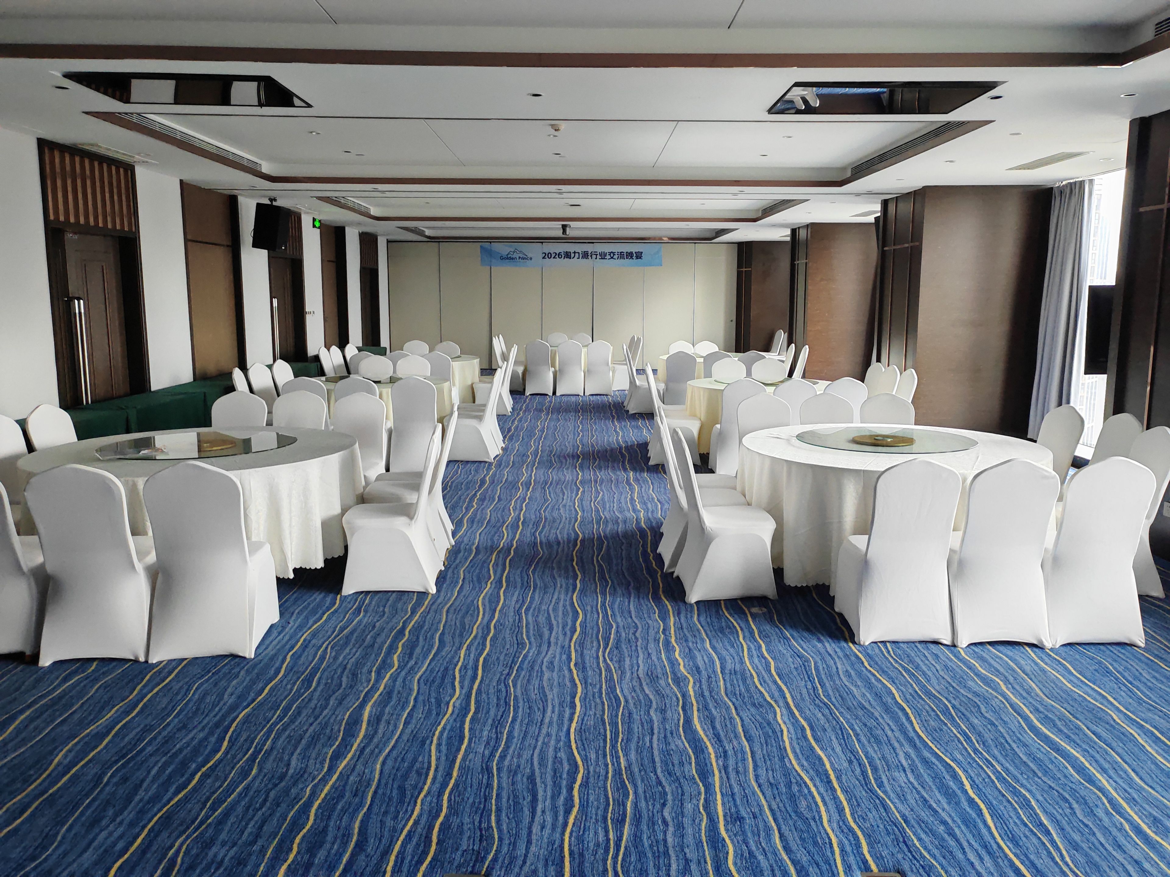 A conference room with round tables and white chairs, a blue carpet, and a sign indicating a future event.