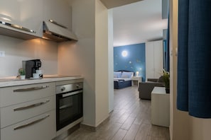 Eine moderne Küche mit Kaffeemaschine, Backofen und Schränken. Ein Schlafzimmer mit Bett, Nachttisch und blauer Wand mit Mondmotiv. Ein Wohnbereich mit Sofa und Pflanze ist sichtbar.