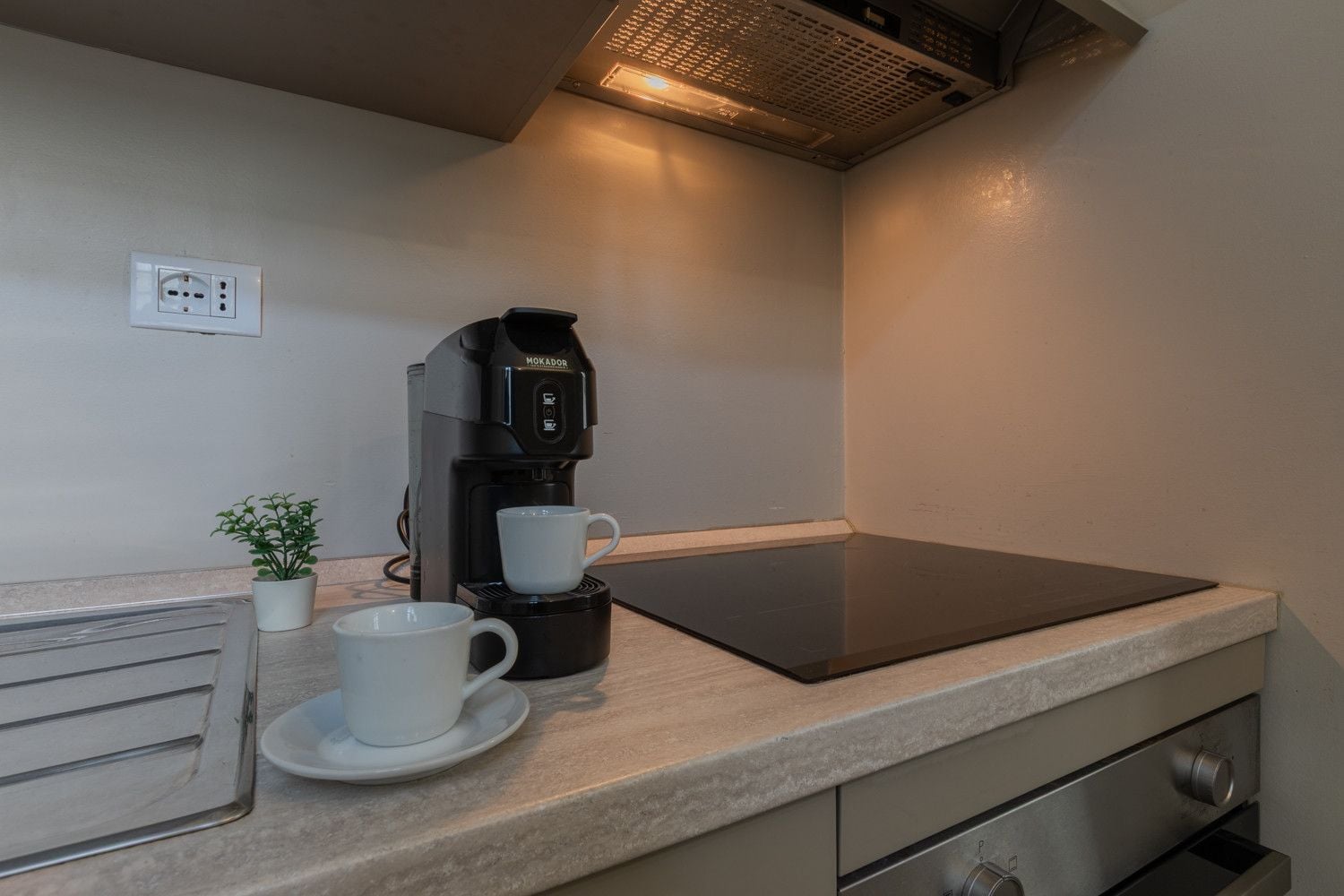 Eine Küchenzeile mit einer Kaffeemaschine, zwei Tassen, einer kleinen Pflanze und einer Steckdose an der Wand.