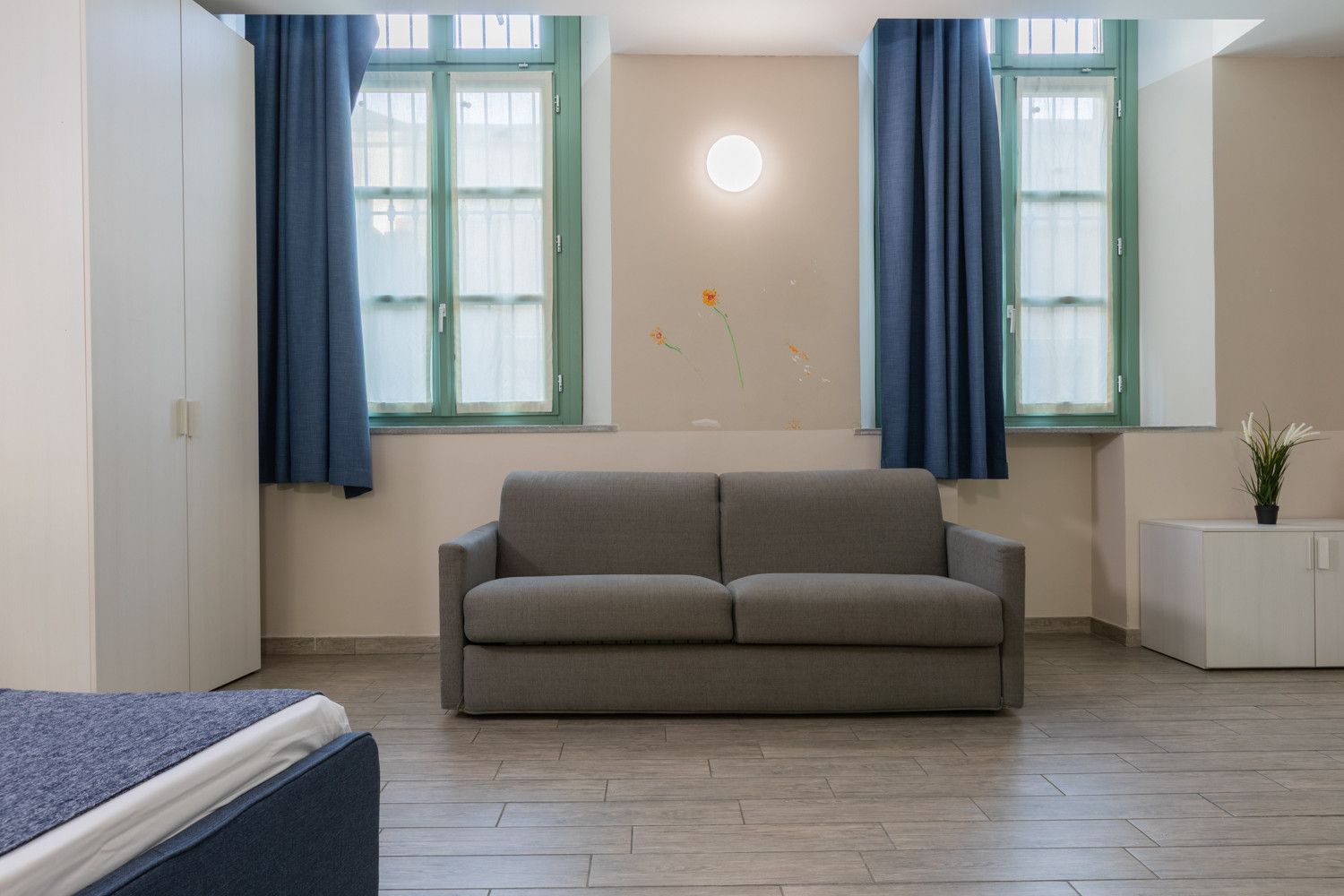 Ein Zimmer mit einem grauen Sofa, zwei Fenstern mit blauen Vorhängen, einem kleinen weißen Schrank und einer wandmontierten Leuchte.