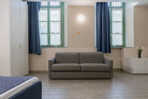 A room with a grey sofa, two windows with blue curtains, a small white cabinet, and a wall-mounted light fixture. - Housing Giulia - Apartment 8 by Wonderful Italy (Turin)