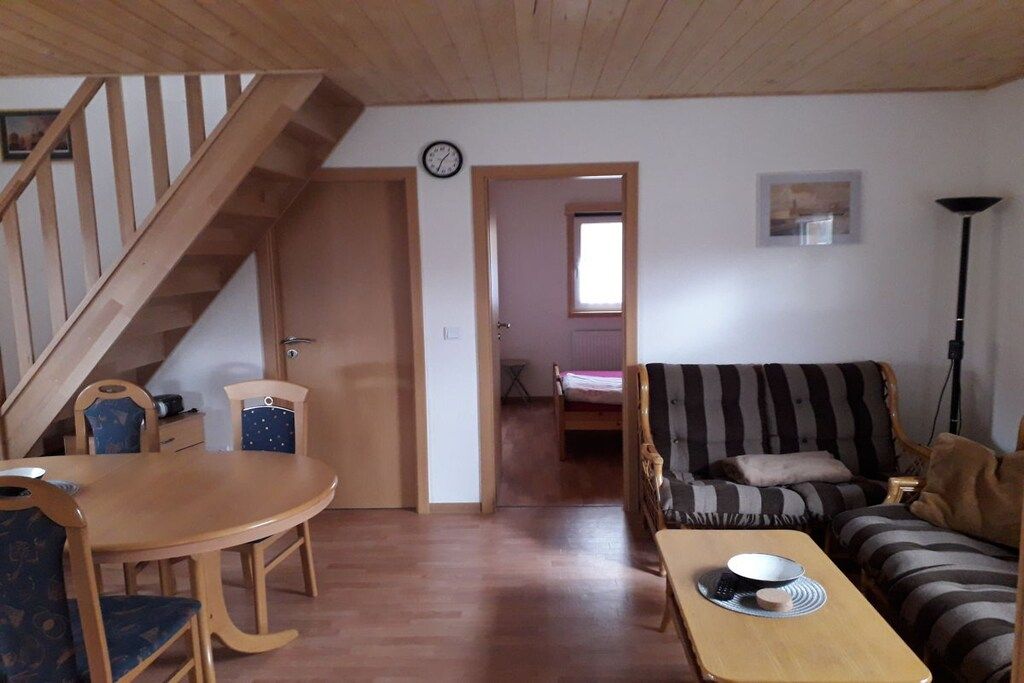 Een kamer met een houten trap, een eettafel met stoelen, een gestreepte bank en een bed op de achtergrond.
