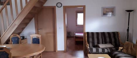 Een kamer met een houten trap, een eettafel met stoelen, een gestreepte bank en een bed op de achtergrond.