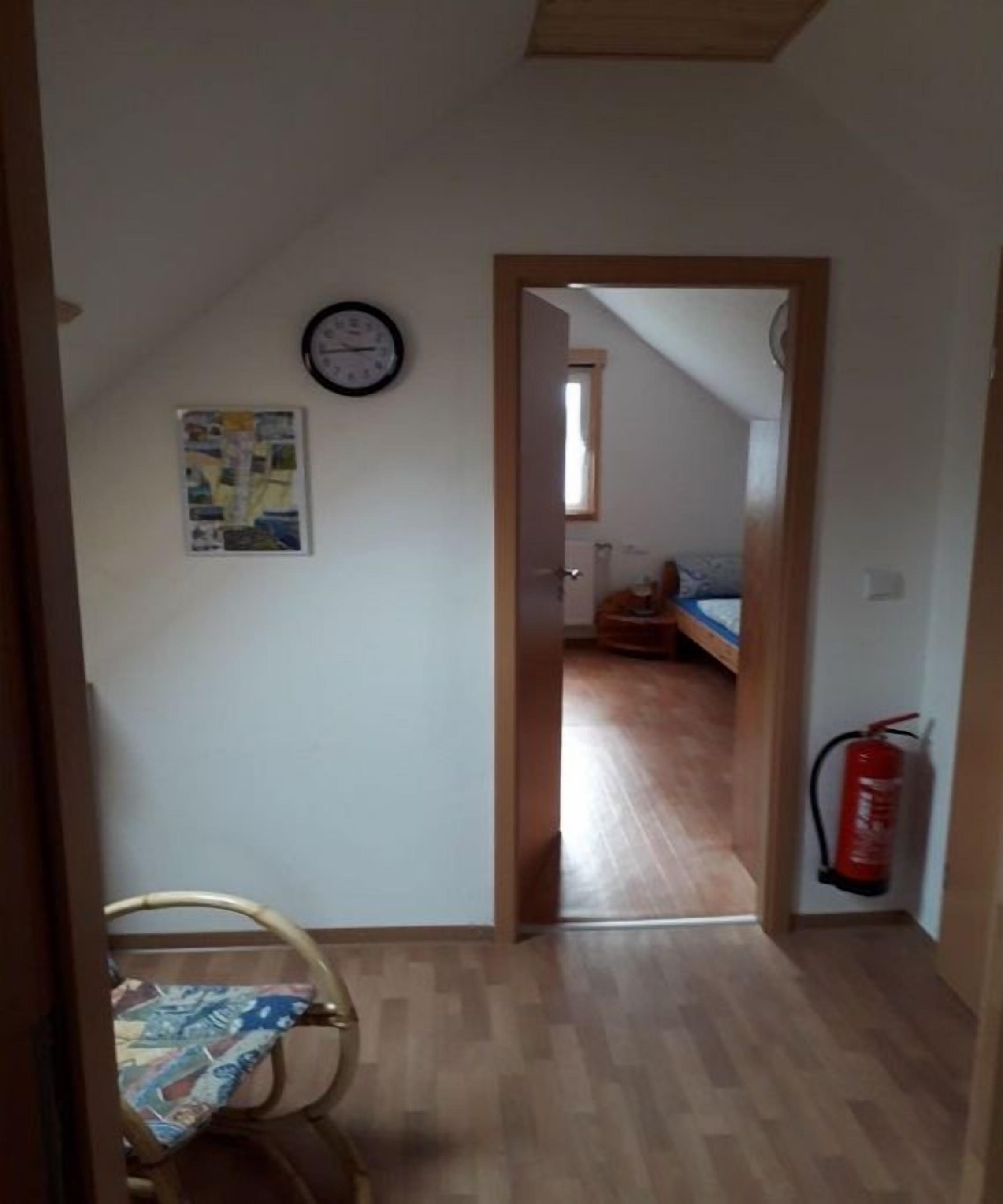 Ein schmaler Flur mit einer Holztür, die in ein Schlafzimmer mit Bett und Fenster führt. Ein Feuerlöscher ist an der Wand angebracht.