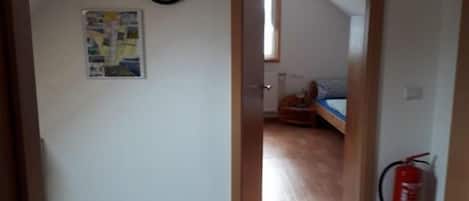 Ein schmaler Flur mit einer Holztür, die in ein Schlafzimmer mit Bett und Fenster führt. Ein Feuerlöscher ist an der Wand angebracht.