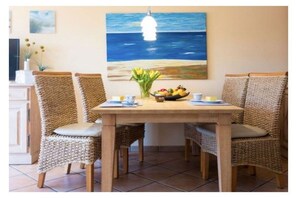 A dining area with a wooden table and wicker chairs, a painting of the ocean on the wall, and a vase with flowers and fruit on the table. - Holiday apartment in Lügde with swimming pool (Lügde)
