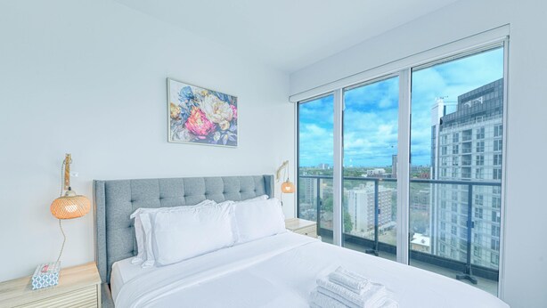 Une chambre d’hôtel moderne avec un grand lit, une vue sur la ville, un tableau au mur et une table de chevet avec une lampe.