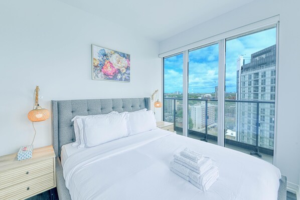 Une chambre d’hôtel moderne avec un grand lit, une vue sur la ville, un tableau au mur et une table de chevet avec une lampe.