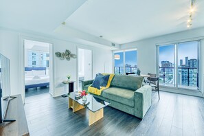 Un salon moderne avec un canapé vert, une table basse en verre et une vue sur la ville grâce à de grandes fenêtres.