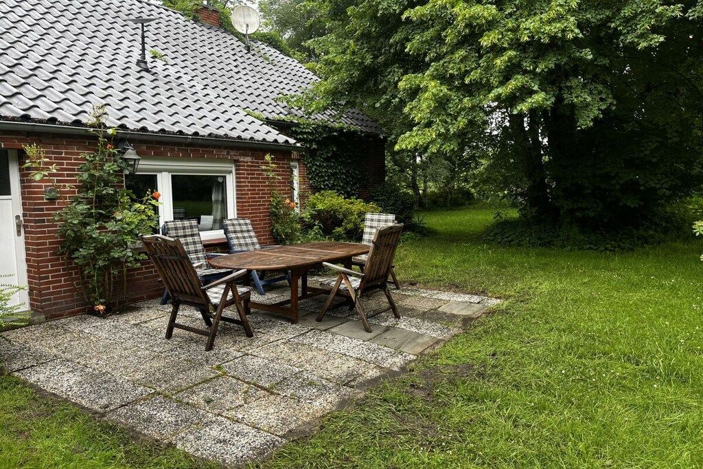 Et murstenshus med et tegltag, en terrasse med bord og stole, samt en have med træer.