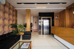 A reception area with a black sofa, a coffee table with magazines, a wooden reception desk, a pillar with a decorative pattern, and an elevator. - Hotel Kiran Inn (Indore)