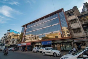 A multi-story building with a glass facade, adjacent shops, and parked cars on the street. - Hotel Kiran Inn (Indore)