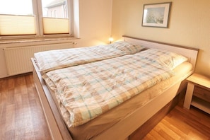 Een eenpersoonsbed met een geruit sprei, een nachtkastje en een raam met jaloezieën.