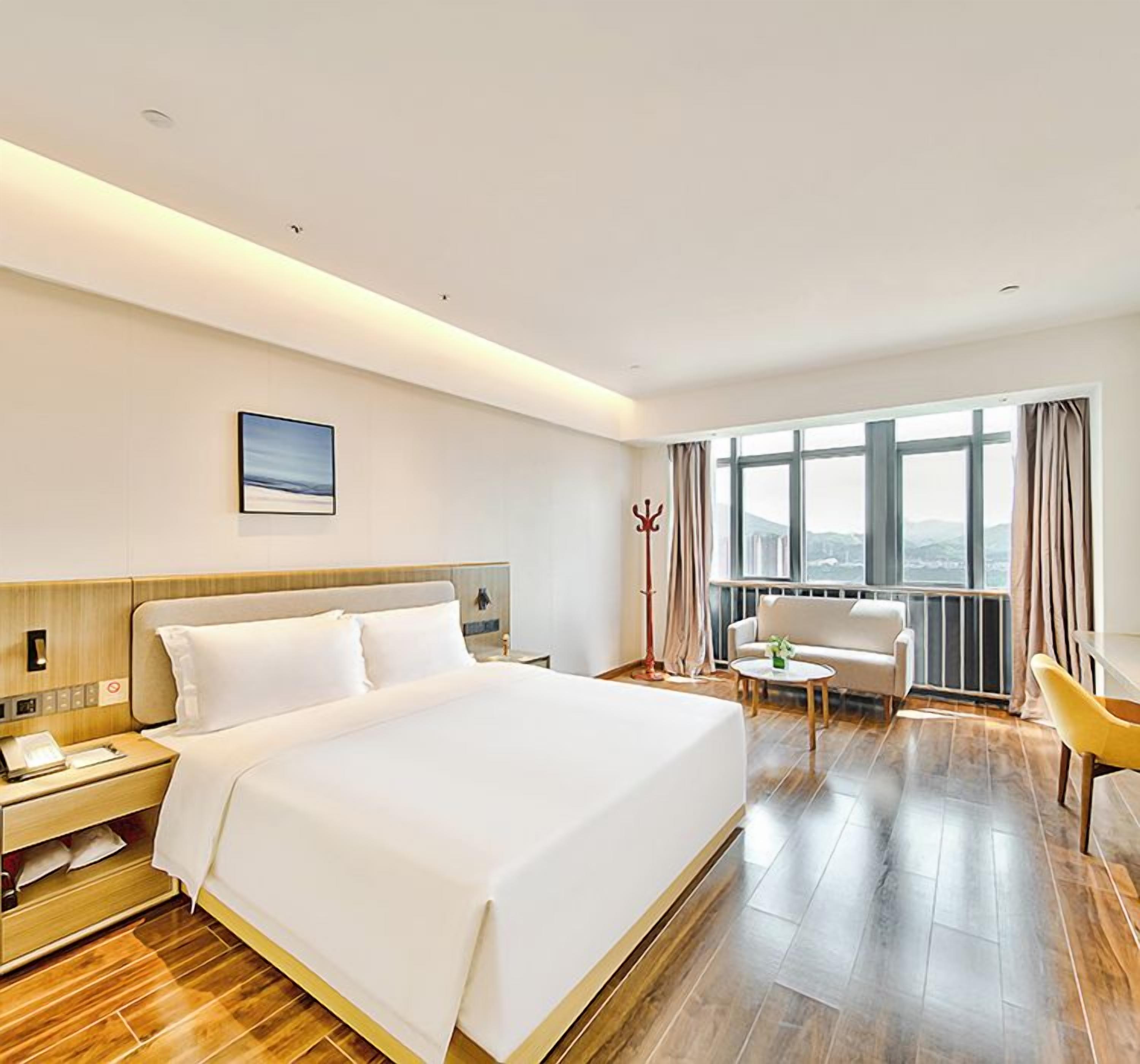 Ein Hotelzimmer mit einem großen Bett, Nachttisch, Lampe und Blick auf die Stadt durch das Fenster.