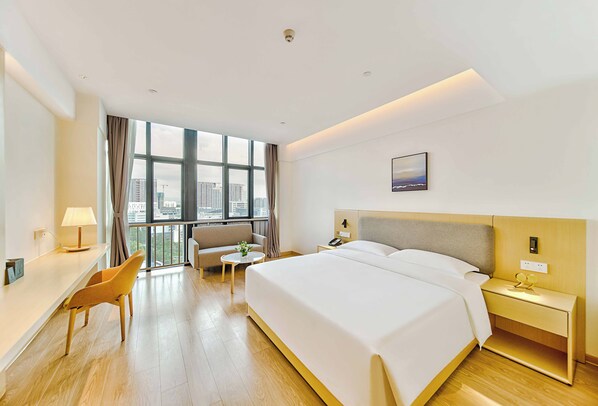 A hotel room with a large bed, a desk, a chair, a small table, and a view of the city through large windows.