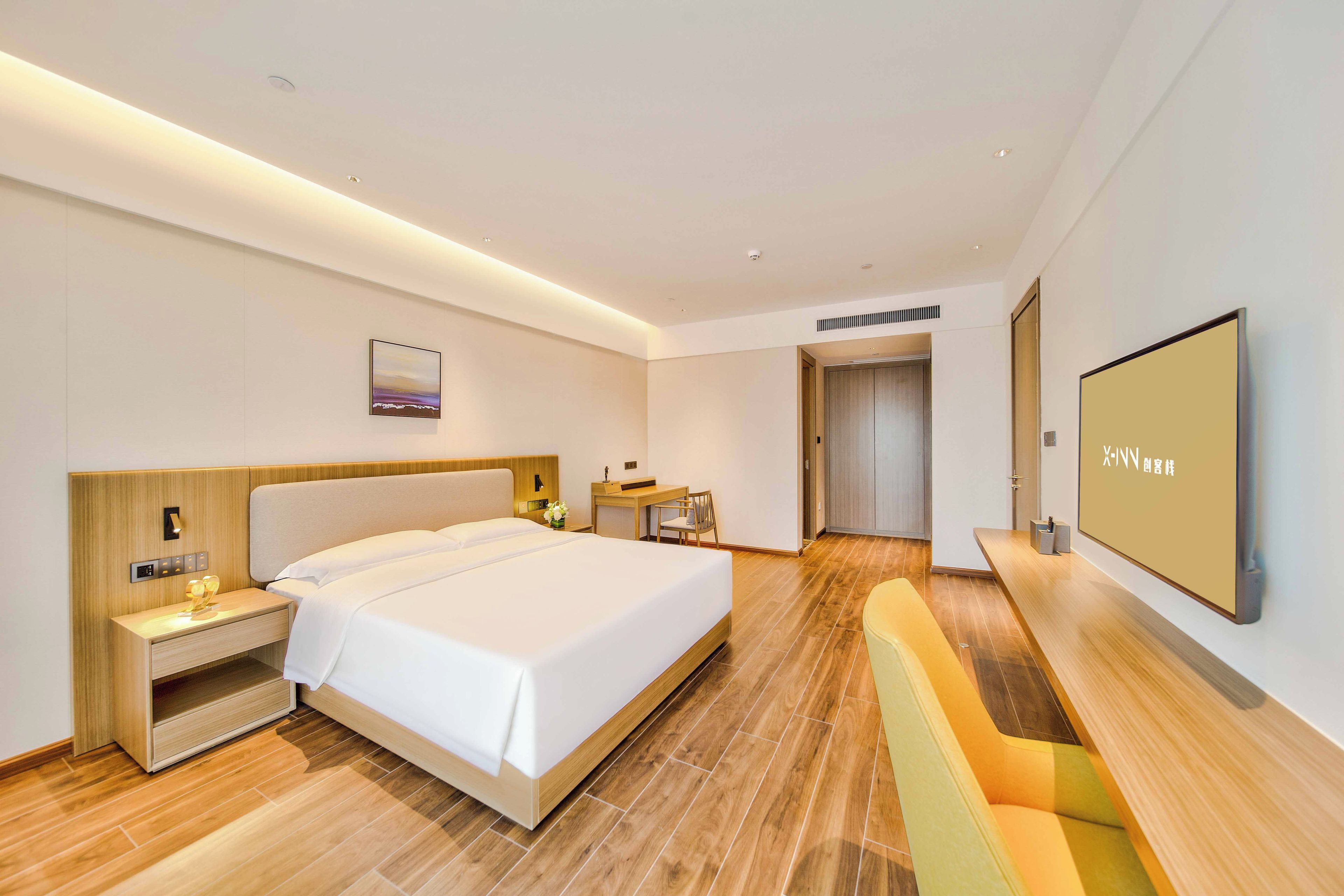 A modern hotel room with a large bed, bedside tables, a desk, a chair, a flat-screen TV, and a door leading to a bathroom.
