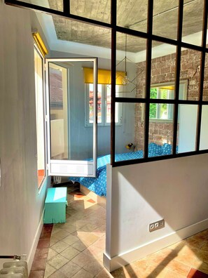 Una habitación con un ventanal grande, una cama con colcha azul estampada y piso de baldosas.