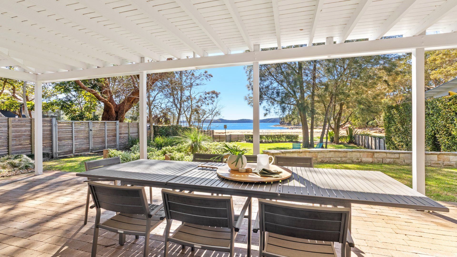 A covered patio with a table and chairs, overlooking a beach and greenery.