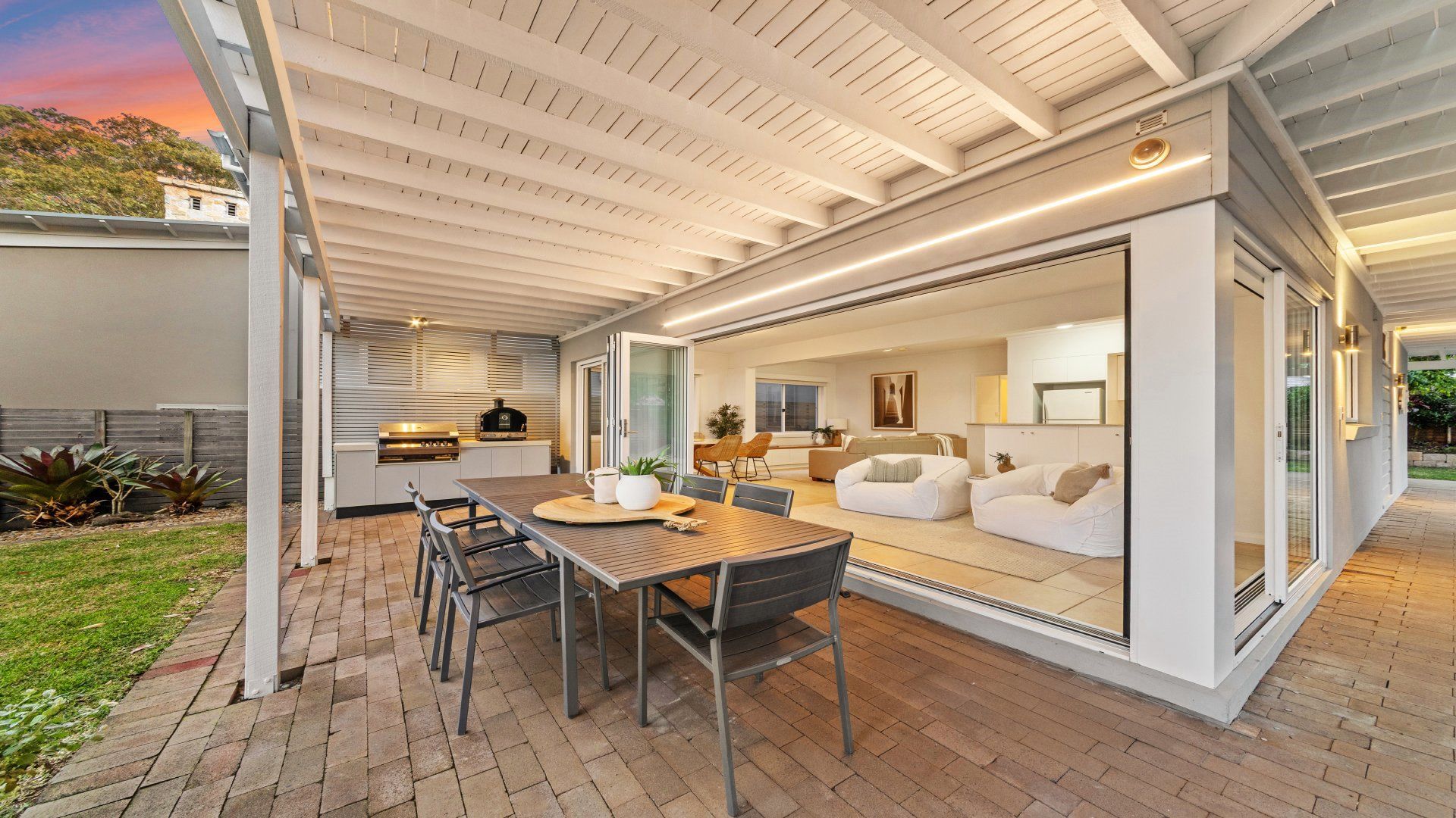 Eine Terrasse im Freien mit Esstisch und Stühlen, einem Grill und einer Glastür, die in einen Wohnbereich mit Sofa und Fernseher führt.