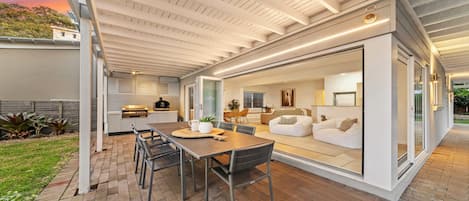 Une terrasse extérieure avec une table et des chaises, un barbecue et une porte-fenêtre coulissante donnant sur un espace de vie intérieur comprenant un canapé et un téléviseur.