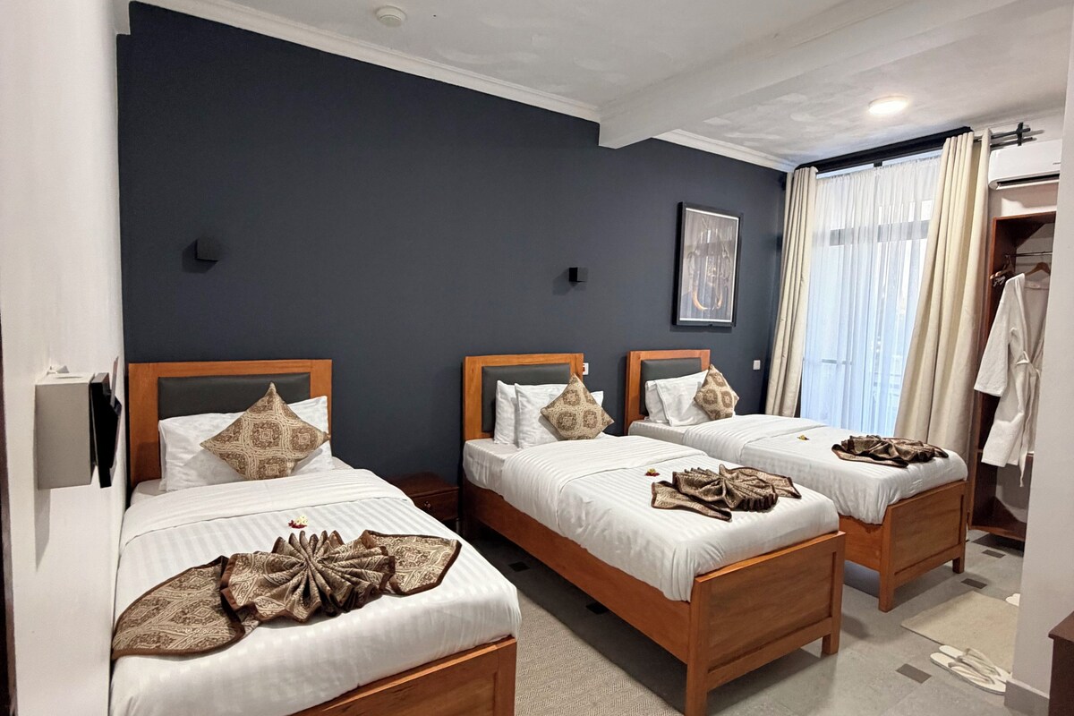 A hotel room with three wooden beds, a large window with curtains, and a wardrobe.