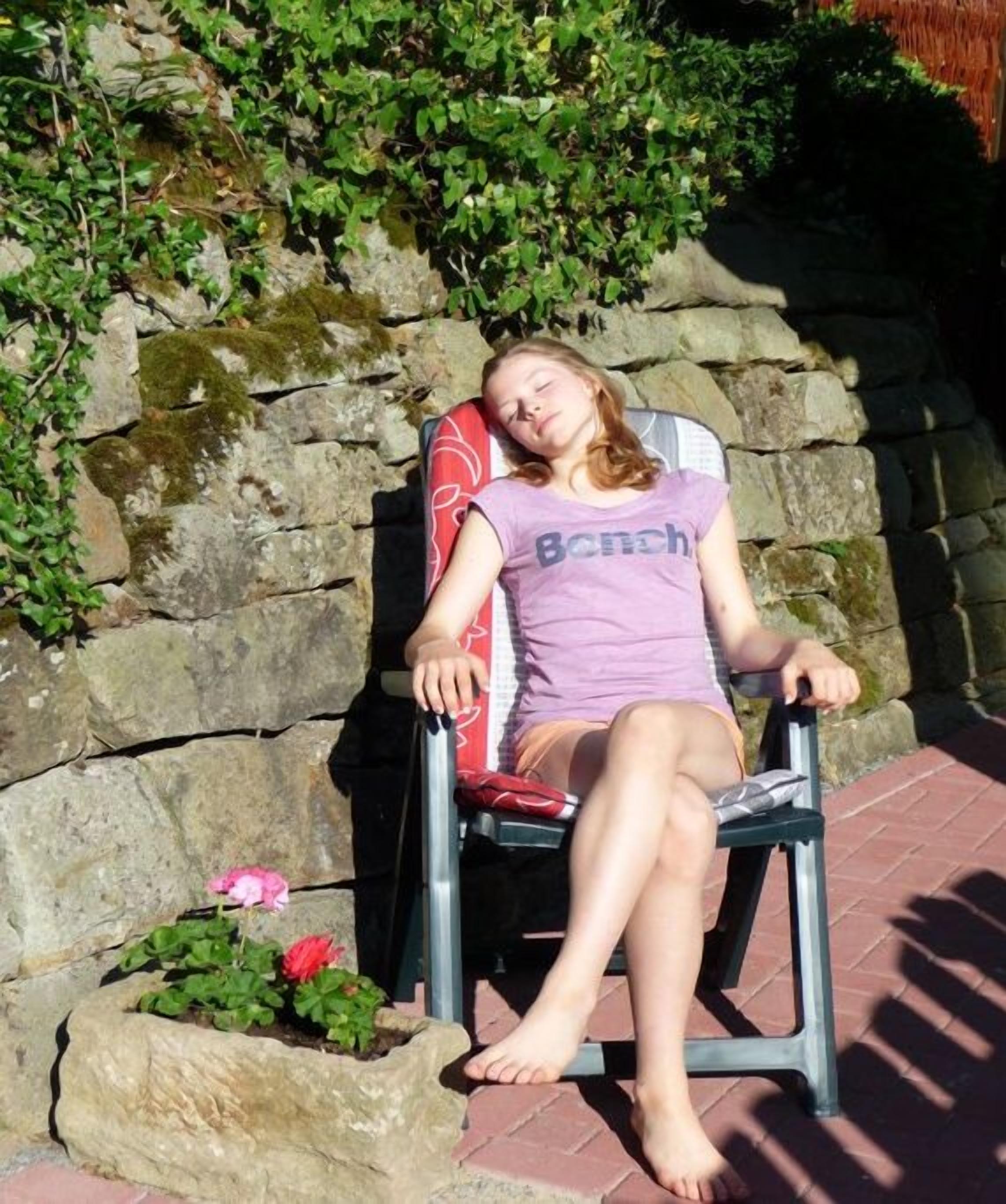 A person sitting on a chair outdoors with a stone wall and greenery in the background.